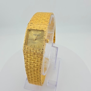May include: A gold-tone Seiko quartz watch with a rectangular face and a textured, woven-style metal link band. The watch dial is gold with thin hour markers, gold hands, and the text Seiko and Water Resistant Quartz printed on the face.