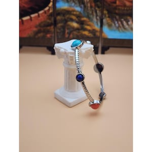 May include: A silver-toned bangle bracelet with turquoise, blue, and coral-colored gemstone accents. The bracelet is displayed on a white pedestal against a neutral background. The gemstones are round and set into the bracelet.