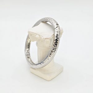 May include: A silver-colored bangle bracelet with a textured, hammered design. The bracelet is hinged and appears to be made of metal. The jewelry is displayed on a white pedestal against a white background.