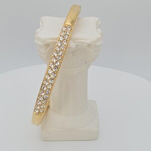 Könnte beinhalten: Ein goldfarbenes Armreif-Armband mit einer Reihe klarer, funkelnder Kristalle. Das Armband wird auf einem weißen Sockel im klassischen Stil präsentiert, der sein elegantes Design hervorhebt. Die Kristalle sind in einem linearen Muster angeordnet.