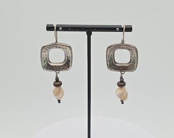 May include: A pair of silver-toned earrings with a square, open-center design. The earrings have a hammered texture and dangle with a small bead and a pearlescent, teardrop-shaped charm. The earrings are displayed on a black stand.