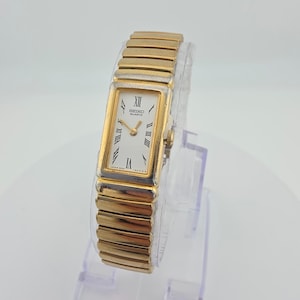 May include: A gold-tone rectangular Seiko Quartz wristwatch with a white dial, black Roman numeral hour markers, and thin black hands. The watch features a flexible, expandable gold-tone metal link bracelet band and a polished gold-tone case.