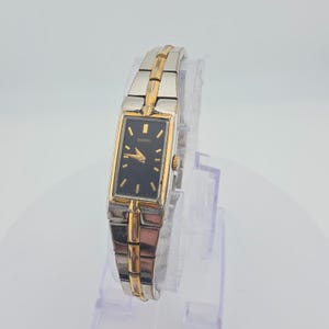 May include: A rectangular Seiko wristwatch featuring a black dial with gold-tone hour markers and hands. The watch has a two-tone metal link bracelet combining silver and gold-tone finishes, with a matching gold-tone bezel and a side winding crown.