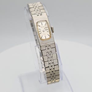Puede incluir: Un reloj de pulsera Seiko vintage con una correa plateada y una esfera rectangular. La esfera del reloj es blanca con detalles dorados y agujas negras. La correa tiene un diseño texturizado. El reloj se muestra sobre un soporte transparente.