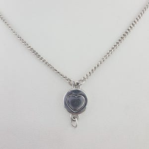 May include: A silver-toned metal necklace featuring a delicate curb chain and a circular pendant. The pendant displays a raised heart shape in the center with a smooth, polished finish. An additional small jump ring hangs from the bottom of the charm.
