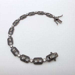 May include: A silver-toned bracelet with a series of rectangular and circular links. Each rectangular link is adorned with small, dark stones and clear, round gemstones. The bracelet is fastened with a clasp.