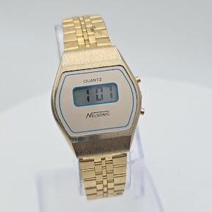 May include: A gold digital wristwatch with a rectangular face. The watch face displays the time, with the word "QUARTZ" above the display and "NELSONIC" below. The watch has a gold-coloured metal strap.