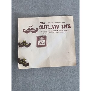 May include: Two brown mustache-shaped pins with yellow text reading Outlaw Inn, displayed on a white paper flyer featuring the text The Outlaw Inn, Kalispell's Convention Center, featuring Hennessy's Steak House, 1701 Hwy. 93 South, Kalispell, Montana 59901.
