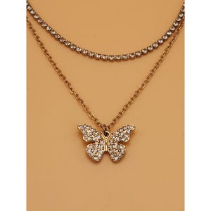 Könnte beinhalten: Eine mehrlagige goldfarbene Halskette mit zwei verschiedenen Ketten. Die obere Kette besteht aus einer feinen Reihe klarer Strasssteine, während die untere Kette ein einfaches Gliederkettendesign mit einem Schmetterlingsanhänger aus Strass ist.