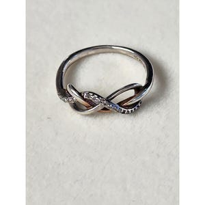 May include: A silver-toned ring with an infinity symbol design. The ring features a twisted band with a small section of the band encrusted with small, clear stones. The center of the infinity symbol has a gold-toned accent.