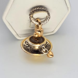 Puede incluir: Un charm de oro y plata con forma de taza de café y platillo. La taza contiene un líquido oscuro, y el platillo tiene la palabra "WAS". Un pequeño charm de corazón dorado cuelga de la taza.