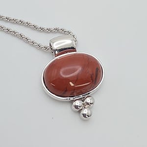 May include: A silver necklace featuring a pendant with a large, oval, reddish-brown stone. The pendant has a silver frame and three small, round silver accents below the stone. The chain is a silver rope style.