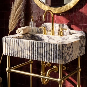 May include: A white and grey marble sink with a fluted exterior design, supported by a gold metal frame. The sink features a high-arched gold faucet, two gold handles, a matching soap dish, and a clear glass soap dispenser with a gold pump.