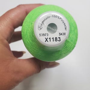 May include: A spool of bright neon green embroidery thread. The top label is white with black text that reads Exquisite, 100% Polyester, 50973, 5KM, and X1183. The thread is wound tightly around the cylindrical spool, showing a vibrant color.