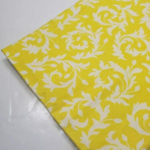 May include: A folded yellow fabric with a white floral pattern. The fabric is a bright yellow color with a repeating design of white, stylized leaves and swirls. The fabric is folded at an angle.