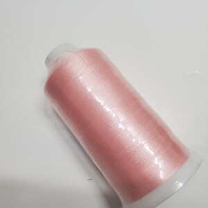 May include: A spool of light pink sewing thread wrapped around a clear plastic cylinder. The thread has a smooth, slightly reflective finish and is wound tightly in a uniform pattern from the top to the bottom of the spool.