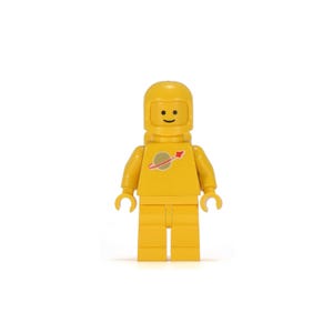 Op de afbeelding: Een gele Lego astronaut figuur met een lachend gezicht en een planeet logo met een rode pijl. De astronaut draagt een helm en een ruimtepak. De figuur staat rechtop.