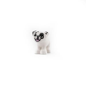 Op de afbeelding: Een wit speelgoedhondje met zwarte vlekken rond de ogen en oren, en een zwarte neus. De hond heeft een glimlachende uitdrukking met een roze tong. Het speelgoed staat op vier witte poten.