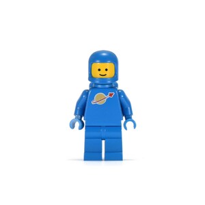 Op de afbeelding: Een blauwe Lego astronaut figuur met een gele gezicht en een blauwe helm. De torso van de astronaut heeft een gouden planeet logo met een rode pijl. De figuur staat rechtop tegen een witte achtergrond.