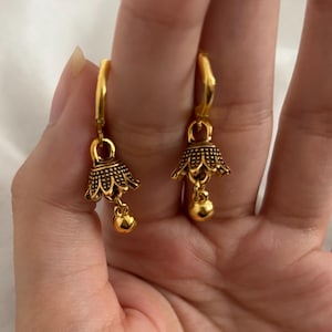 May include: A pair of gold-coloured earrings. Each earring features a bell-shaped design with intricate detailing and a small, round charm at the bottom. The earrings are suspended from a gold-coloured hoop.