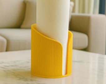 May include: A yellow paper towel holder with a cylindrical design, holding a roll of white paper towels. The holder has vertical lines and sits on a marble-topped table. The background includes a sofa and framed artwork.