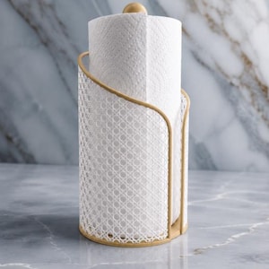 Könnte beinhalten: Eine weiße Küchenrolle in einem dekorativen Halter. Der Halter hat einen goldfarbenen Rahmen und ein weißes, gewebtes Muster. Eine kleine, goldene Kugel bedeckt die Rolle. Der Halter steht auf einer Marmoroberfläche.