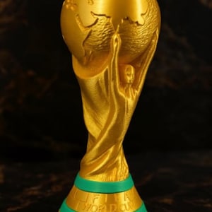 May include: A gold-colored trophy with a globe held up by two figures. The base is green with the words "FIFA WORLD CUP" in gold. The trophy is a symbol of victory and achievement.