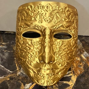 May include: A gold-colored mask with intricate, raised floral and scrollwork patterns. The mask has eye openings and a nose, mouth, and chin. The mask is likely for decorative purposes or costume use.