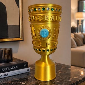 May include: A gold-colored trophy cup featuring the text FUSSBALL and a central circular emblem with a light blue logo. The cup is decorated with alternating green and clear circular gems around the top and bottom rims and two figures.
