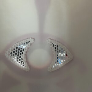 May include: Close-up of a white object with two triangular, mesh-like sections. The mesh has small, dark circular holes. The background is a gradient of light pink and white, with a central, elongated shape.