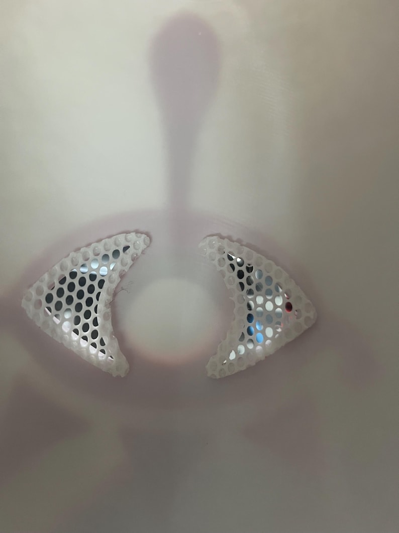 May include: Close-up of a white object with two triangular, mesh-like sections. The mesh has small, dark circular holes. The background is a gradient of light pink and white, with a central, elongated shape.