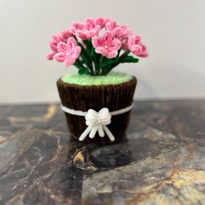May include: A handmade floral decoration in a cupcake-shaped pot. The pot is brown with a white bow, and the arrangement features pink flowers with green stems and leaves. The arrangement sits on a marble surface.