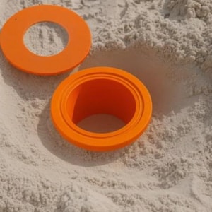 May include: Two bright orange plastic rings, one with a raised edge, rest on light-colored sand. The rings are circular with a hole in the center. The image is taken in natural light.