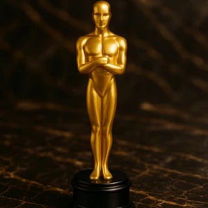 May include: A gold-colored statuette of a man with arms crossed, standing on a black circular base. The figure has a smooth, reflective surface and is set against a dark, patterned background. The statue is a symbol of achievement.