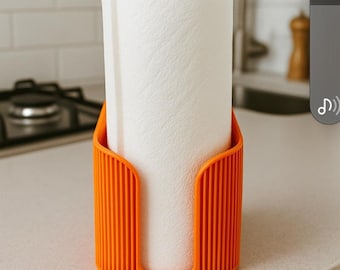 May include: An orange paper towel holder with a roll of white paper towels. The holder has a vertical, ribbed design and is sitting on a light-colored countertop. The kitchen setting includes a stovetop and sink.