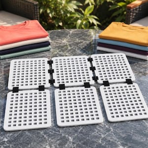 May include: A white, foldable, grid-patterned trivet with black hinges, displayed alongside two stacks of folded t-shirts in various colors including coral, cream, navy, sage green, mustard yellow, light blue, purple, and white on a marble surface.