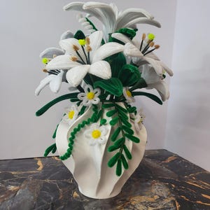 Può includere: Un vaso di ceramica bianca con un design strutturato, riempito di gigli bianchi fatti a mano, foglie verdi e piccole margherite bianche con centri gialli. La composizione è realizzata con una varietà di materiali, creando un'esposizione floreale unica.