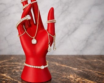 Op de afbeelding: Een rode handvormige sieradenhouder met diverse goudkleurige ringen, een armband, een ketting en oorbellen. De sieraden zijn versierd met heldere edelstenen. De houder staat op een bruin en grijs marmeren oppervlak.