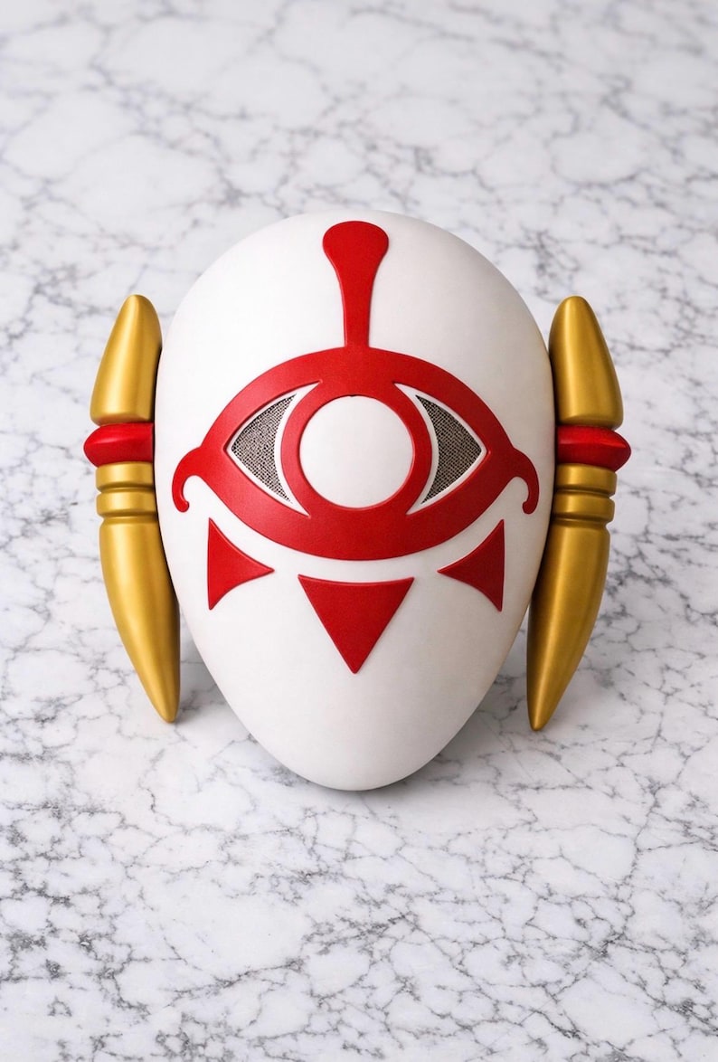 May include: White mask with a red eye design, red accents, and gold side pieces. The mask has a symmetrical design with a central eye symbol and is likely a costume or decorative item.