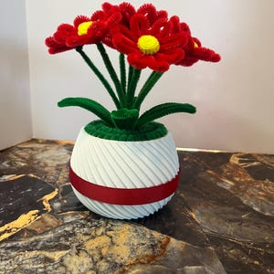 May include: A handmade floral arrangement featuring red pipe cleaner flowers with yellow centers and green stems and leaves. The flowers are in a white, ribbed pot with a red ribbon accent. A unique, decorative piece.