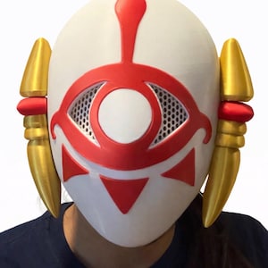 May include: A white mask with a red eye design and gold accents. The mask features a central eye shape with a mesh interior, red triangles, and a red vertical line. Gold, cone-shaped elements are on each side.
