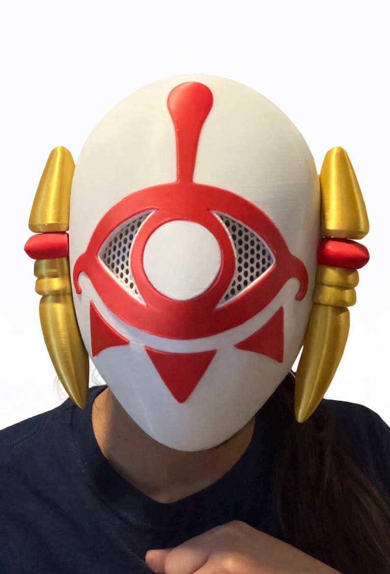 May include: A white mask with a red eye design and gold accents. The mask features a central eye shape with a mesh interior, red triangles, and a red vertical line. Gold, cone-shaped elements are on each side.