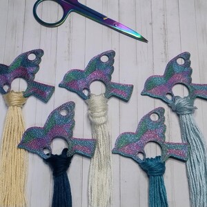 Puede incluir: Un conjunto de organizadores de hilo con forma de pájaro en colores iridiscentes, cada uno con una borla de diferente color. Unas tijeras de arcoíris en la parte superior. Diseñados para proyectos de bordado o punto de cruz.