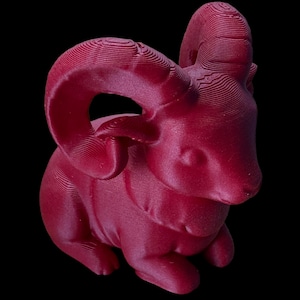 May include: A maroon ram sculpture with large, curved horns and a textured surface. The ram is in a seated position, with its head turned slightly to the side. The background is solid black.