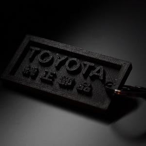 May include: Black rectangular keychain with the word "TOYOTA" in raised letters, along with Japanese characters. The keychain is attached to a metal ring. The background is dark.