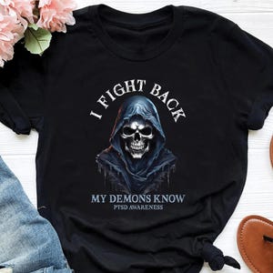 Puede incluir: Camiseta negra con un gráfico de una calavera con capucha. El texto "I FIGHT BACK" está encima de la calavera, y "MY DEMONS KNOW PTSD AWARENESS" está debajo. La camiseta se muestra sobre una superficie blanca.