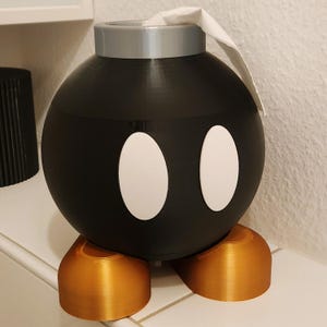 Könnte beinhalten: Ein schwarzer Toilettenpapierhalter in Form einer Cartoon-Bombe mit weißen ovalen Augen und goldfarbenen Füßen. Die Oberseite ist silberfarben, und ein Stück weißes Toilettenpapier ragt heraus.