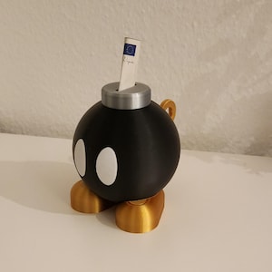 Puede incluir: Una hucha negra, esférica, con forma de bomba de dibujos animados, con ojos ovalados blancos y pies y asa dorados. Un billete de papel se inserta en una ranura en la parte superior. Una forma divertida y original de ahorrar dinero.