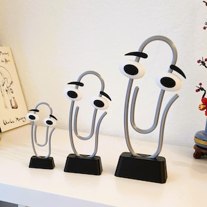 Peut inclure: Trois sculptures fantaisistes en forme de trombone de différentes tailles. Chaque trombone en métal gris est orné de grands yeux de dessin animé avec des pupilles noires et un sourcil noir. Les trombones reposent sur des socles rectangulaires noirs.
