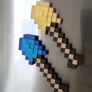Könnte beinhalten: Zwei pixelige, 3D-gedruckte Spitzhacken. Eine hat einen gelben Kopf und einen braunen und beigefarbenen Griff. Die andere hat einen blauen Kopf und einen braunen und beigefarbenen Griff. Wahrscheinlich Magnete oder Dekorationsartikel.